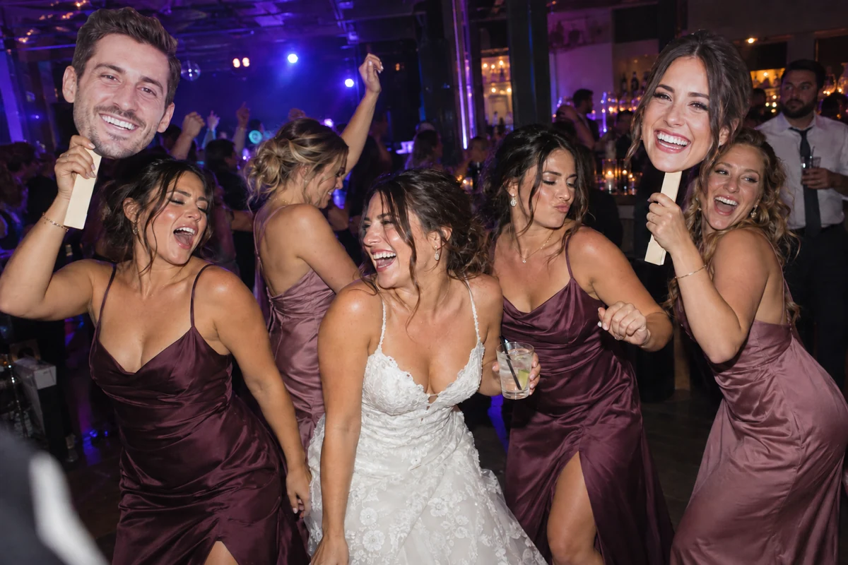 A bride and bridesmaids dancing with bride and groom head signs in a flash-lit, club-style wedding reception